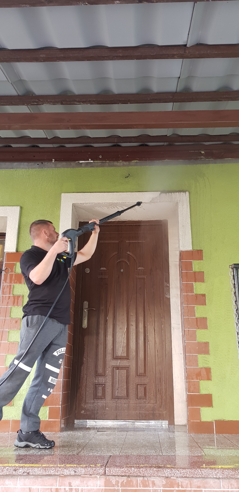 Mężczyzna czyści elewację domu myjką ciśnieniową, widoczny strumień wody i czyszczona ściana z zieloną farbą, brązowe drzwi i mokry chodnik.