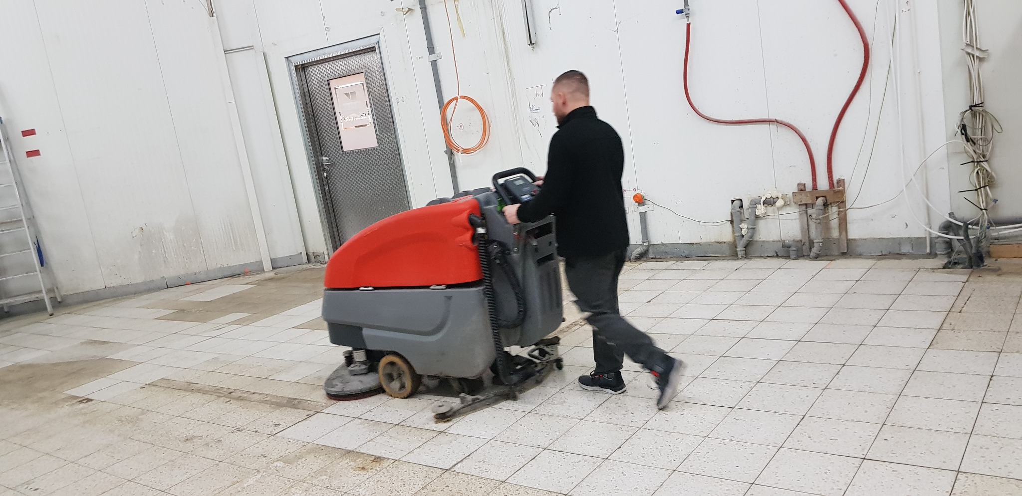 Mężczyzna obsługuje maszynę do czyszczenia podłóg w pomieszczeniu przemysłowym z białymi ścianami i szarą podłogą w kratkę. Widoczne ślady po czyszczeniu.