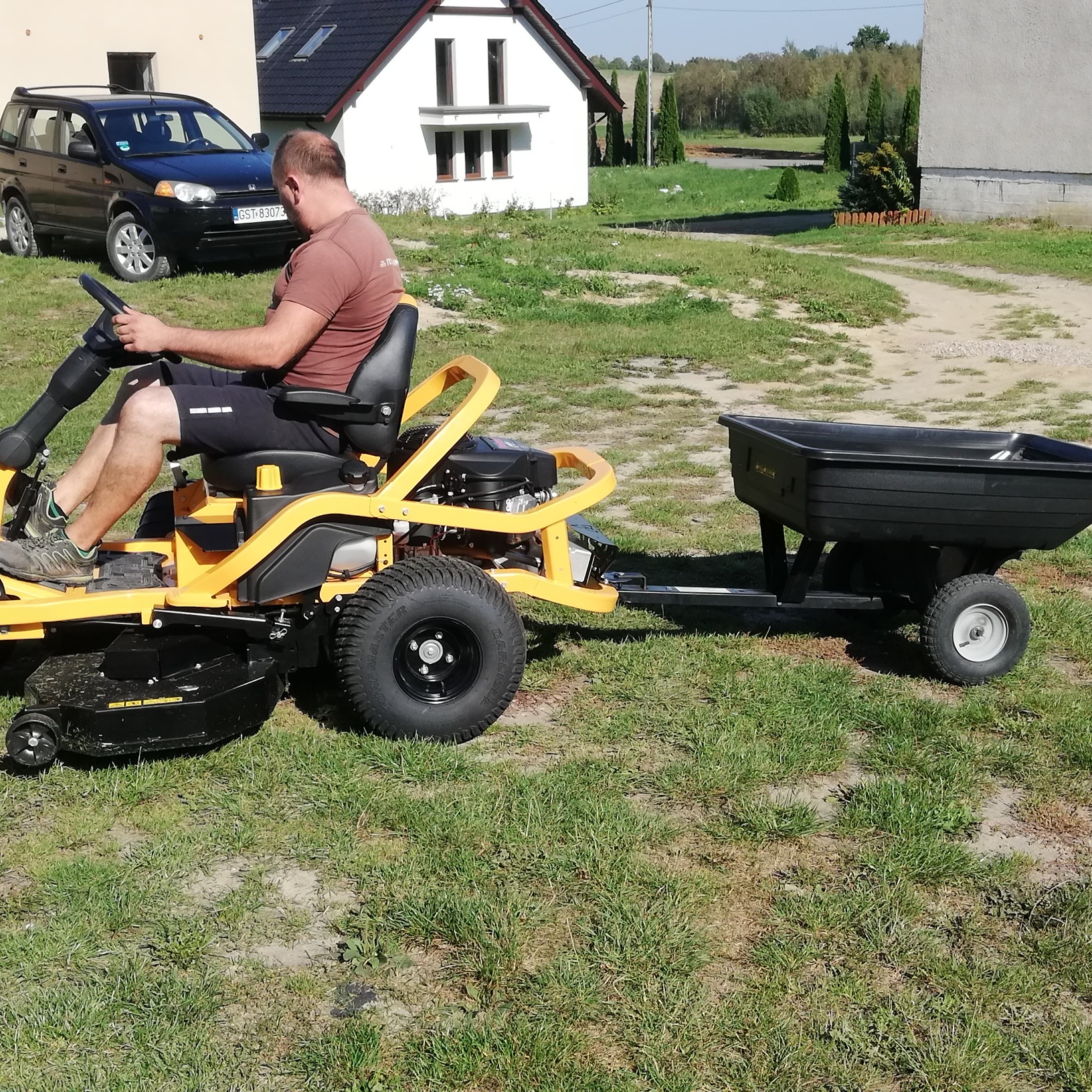 Mężczyzna kosi trawę traktorkiem z przyczepką na tle domu i samochodu.