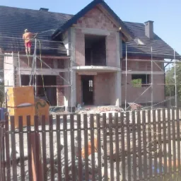 Budowa domu w stanie surowym otwartym, widoczne rusztowania, murarz na dachu układa dachówkę, ogrodzenie placu budowy z drewnianych sztachet.