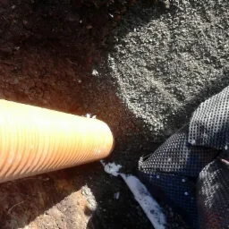 Żółta rura drenarska ułożona w wykopie, częściowo zasypana żwirem, obok czarna membrana kubełkowa.