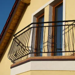 Balkon z czarną, kutą balustradą z ozdobnymi elementami w kształcie liści na tle żółtej elewacji domu z widocznym fragmentem dachu. Balustrada zamontowana na niewielkim balkonie wykończonym płytkami.