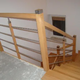 Jasne drewniane schody z balustradą z poziomych metalowych prętów, widok z góry na pierwszym biegu, fragment surowej posadzki i okno z żaluzjami w tle.