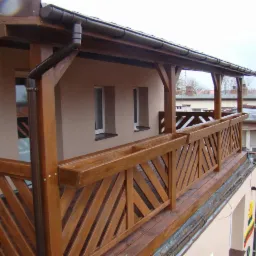 Zadaszony balkon z drewnianą balustradą o ukośnym wzorze, wyposażony w podłużne donice, widok z zewnątrz na budynki w tle.