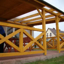 Zadaszona konstrukcja z drewna pomalowana na żółto w Kołobrzegu, z widocznym fragmentem dachu z przezroczystego tworzywa i pracującym mężczyzną w tle, na tle domu i ułożonego drewna.