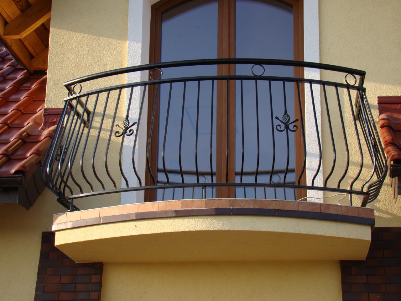 Czarna, metalowa balustrada balkonowa z ozdobnymi elementami na tle okna i elewacji budynku.
