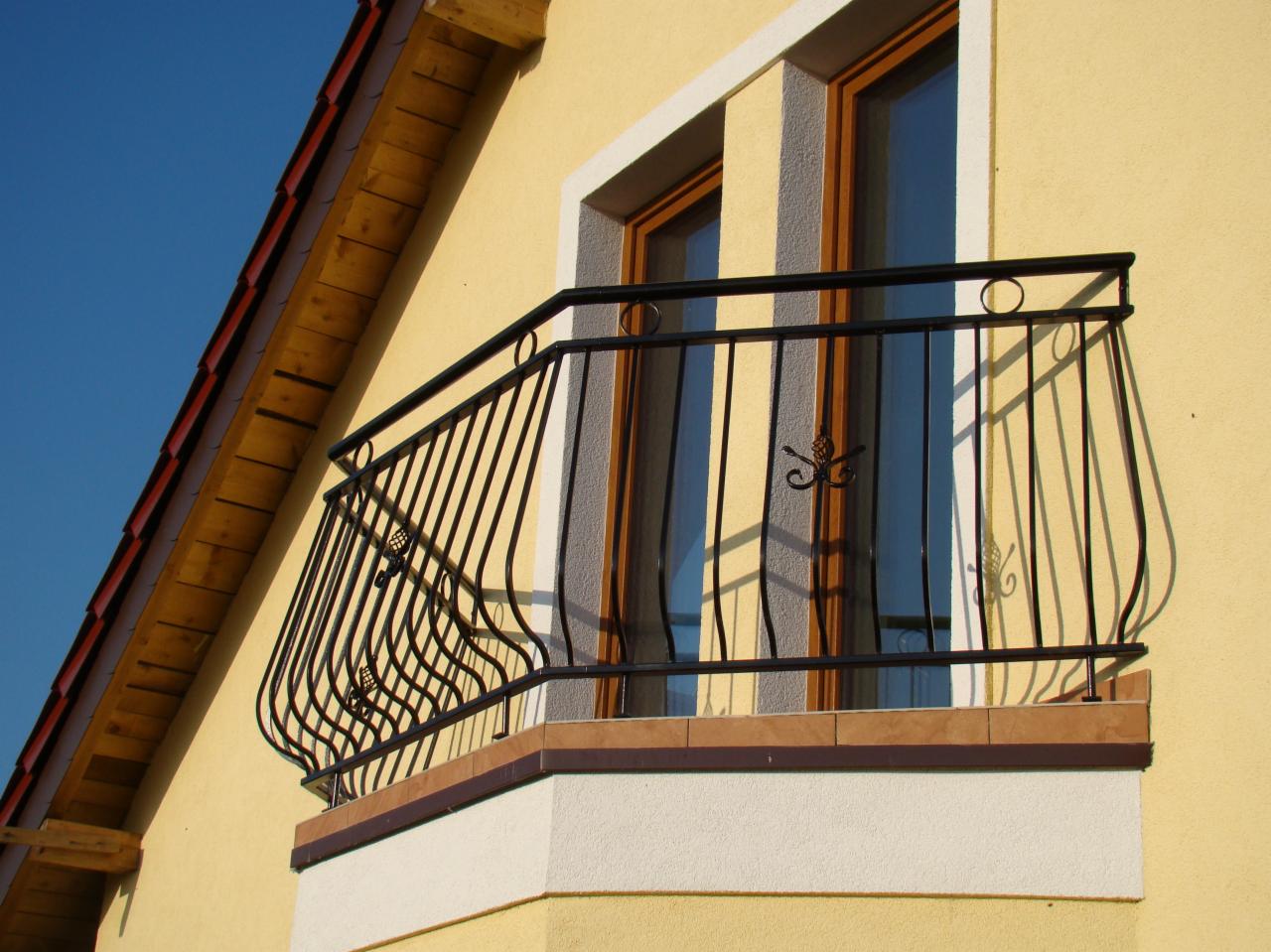 Balkon z czarną, kutą balustradą z ozdobnymi elementami w kształcie liści na tle żółtej elewacji domu z widocznym fragmentem dachu. Balustrada zamontowana na niewielkim balkonie wykończonym płytkami.