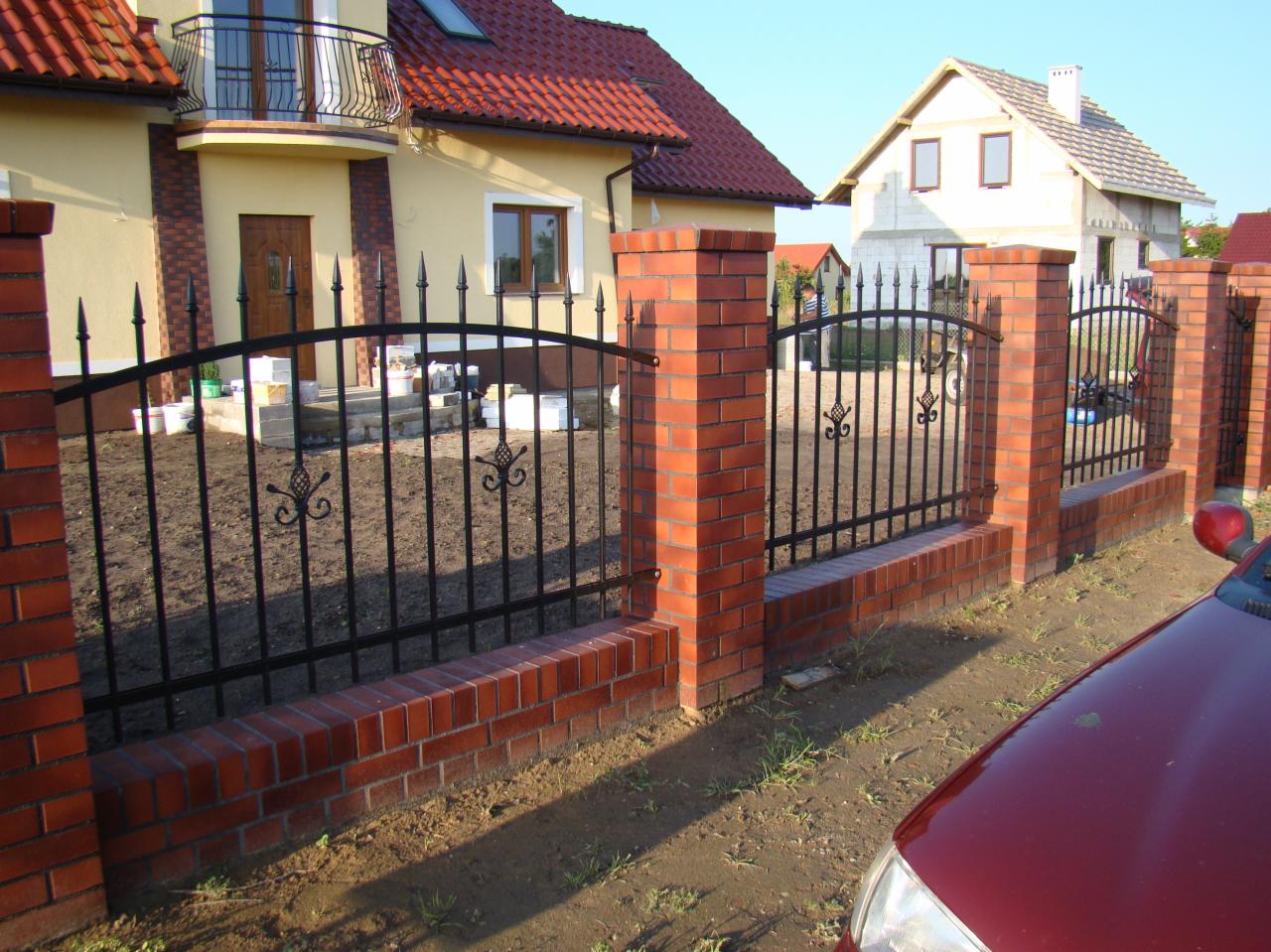 Nowoczesne ogrodzenie z czarnych, kutych przęseł i ceglanych słupków, otaczające posesję z domem w tle.