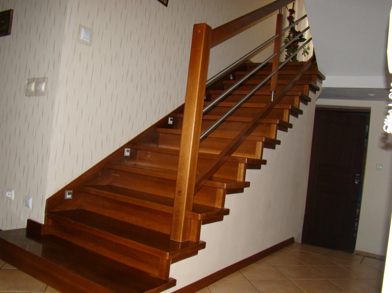 Klatka schodowa z drewna o ciepłym odcieniu, z metalową balustradą i subtelnym oświetleniem punktowym w stopniach, prowadząca do ciemnych drzwi.