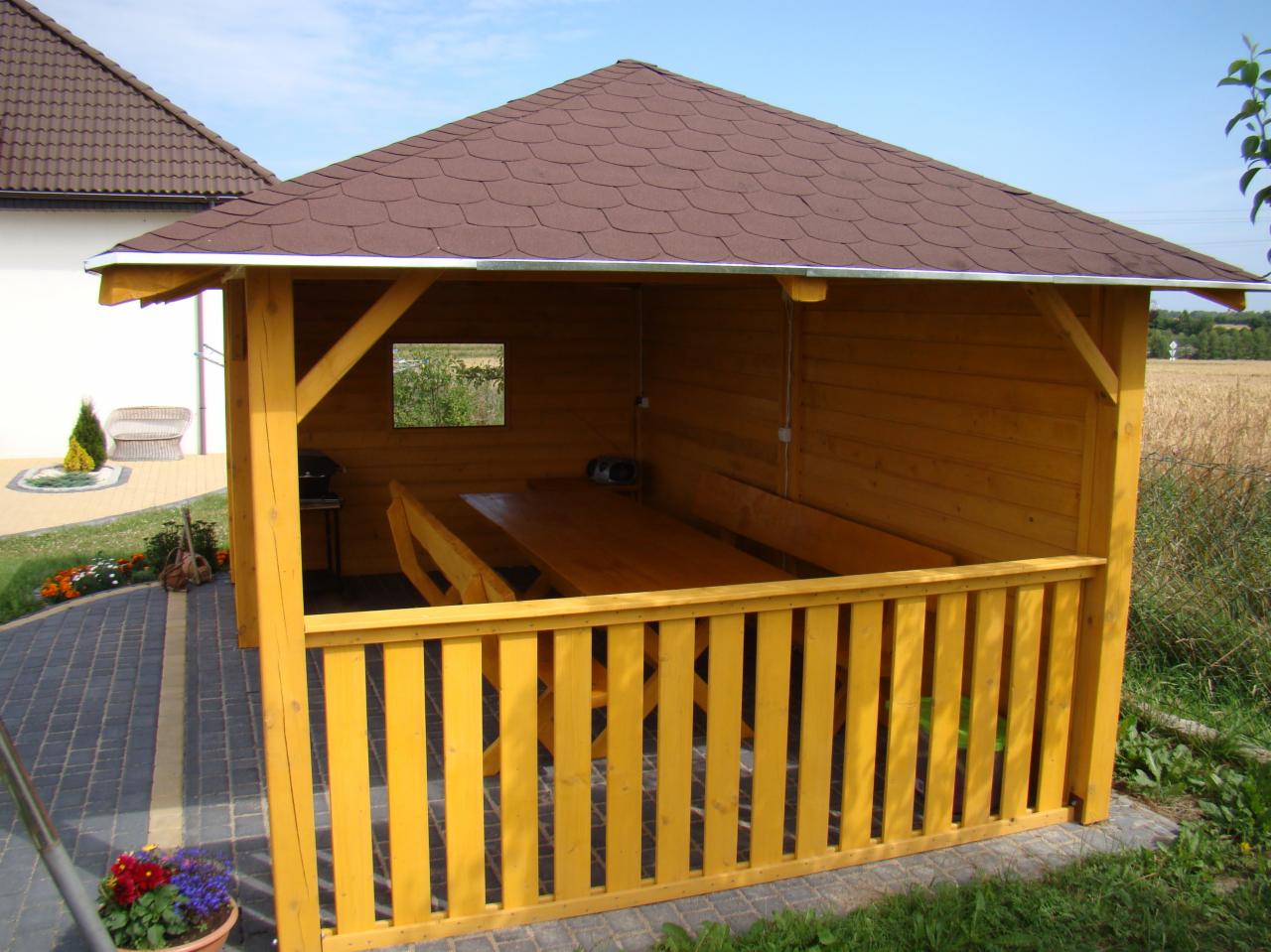 Żółta, drewniana konstrukcja z brązowym dachem, pełniąca funkcję zadaszonego miejsca do siedzenia z drewnianym stołem i ławkami wewnątrz, widok z zewnątrz, z fragmentem ogrodu i domu w tle.