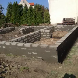 Wznoszenie fundamentów domu z bloczków betonowych, widoczne zbrojenie i izolacja przeciwwilgociowa, teren przygotowany pod dalsze prace budowlane.