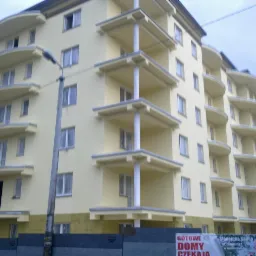 Żółty budynek wielorodzinny z balkonami w trakcie prac wykończeniowych, widoczny pracownik na jednym z balkonów i baner reklamowy u dołu.