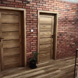 Dwa drewniane panele drzwiowe z poziomymi wstawkami na tle ściany z cegły dekoracyjnej, uzupełnionej drewnopodobną podłogą i małą rośliną doniczkową.