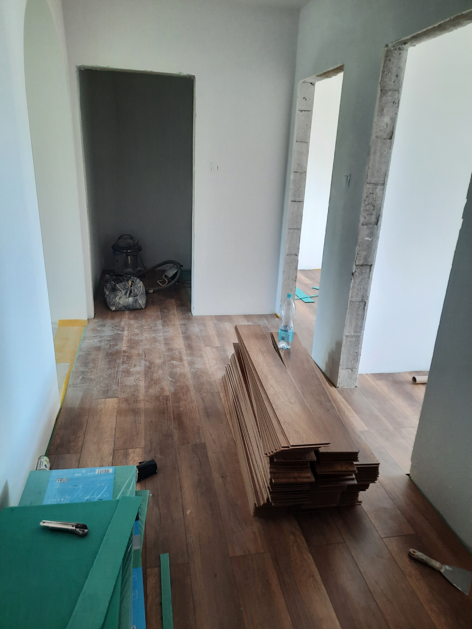 Stos brązowych paneli podłogowych ułożony na podłodze w trakcie remontu, widoczny nóż do tapet, szpachelka, butelka wody i worki z narzędziami w tle.