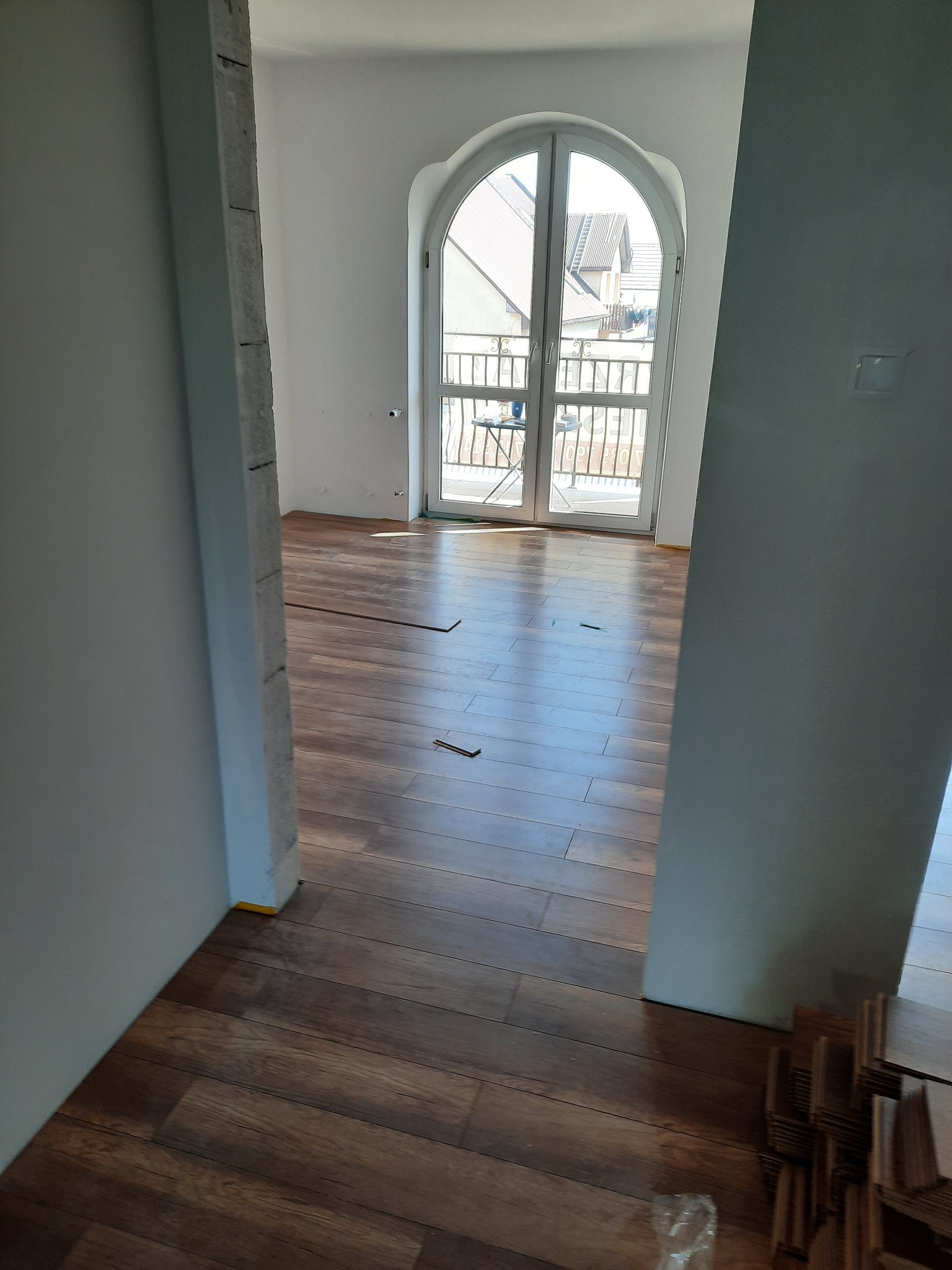 Układanie paneli podłogowych w pokoju z dużym oknem balkonowym, widoczne fragmenty paneli i kolumna w trakcie wykończenia.