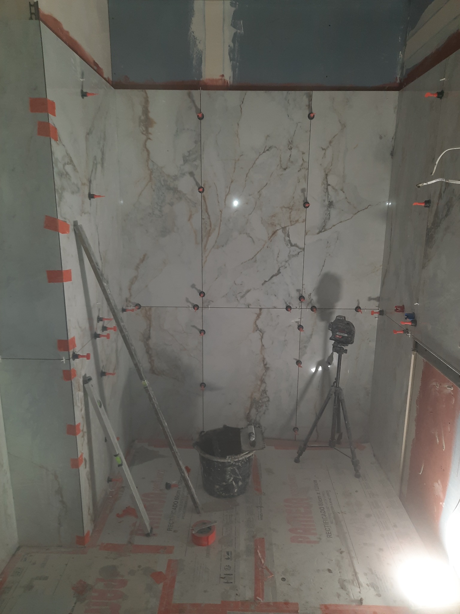 Montaż marmurowych płytek w łazience z użyciem poziomicy laserowej i systemu poziomowania płytek. Widoczny kubeł z zaprawą, poziomica i zabezpieczona folią podłoga.