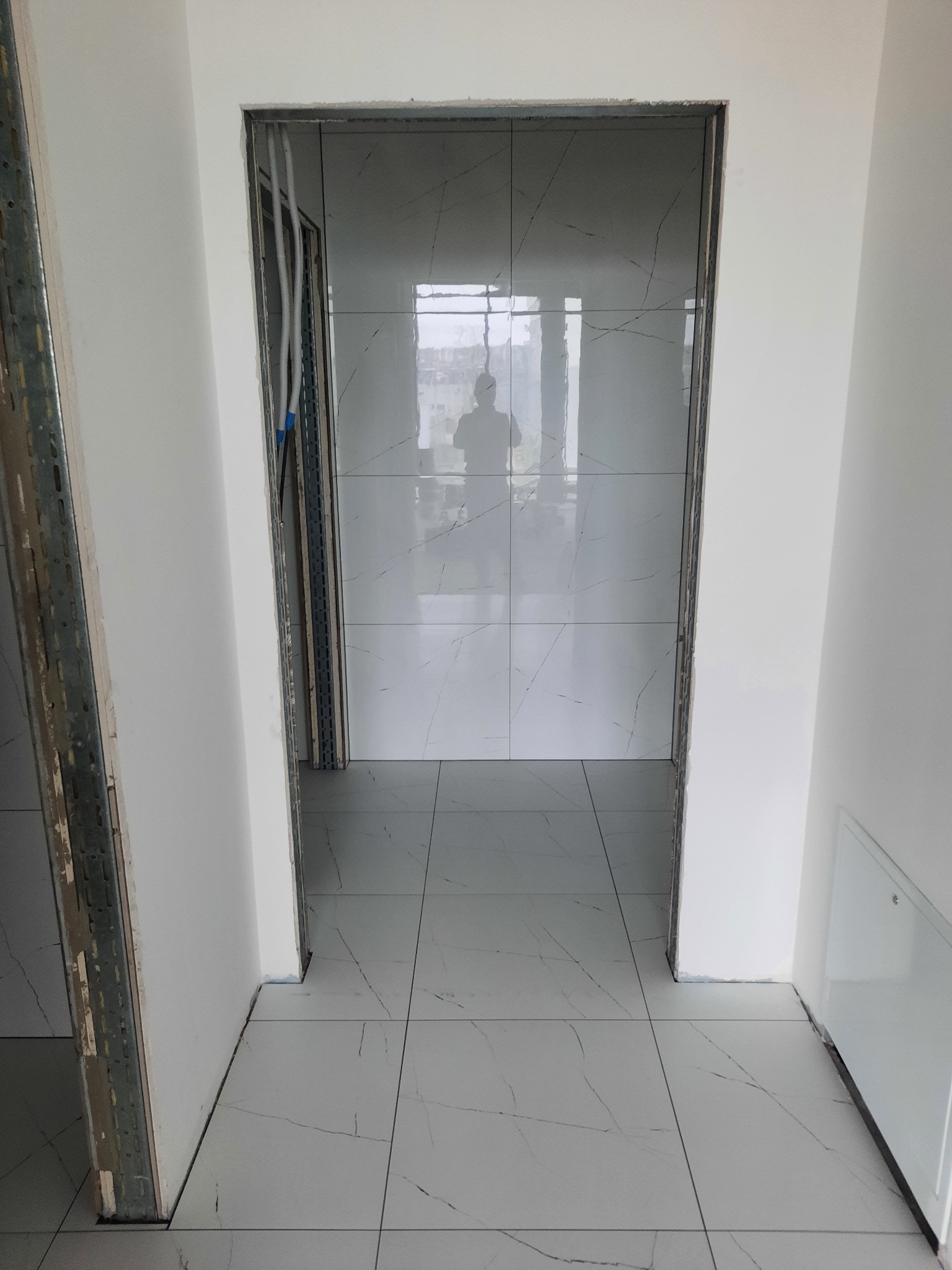 Wnętrze w trakcie remontu z ułożoną jasną podłogą z płytek imitujących marmur, widoczne profile metalowe przy ścianie i instalacje w suficie, odbicie postaci w lustrzanej powierzchni płytek.