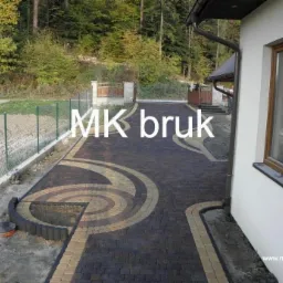 Ułożona ścieżka z ciemnej kostki brukowej z jasnymi, zakrzywionymi wzorami, obok białego budynku z brązowym oknem i siatki ogrodzeniowej.