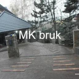 Ułożony podjazd z kostki brukowej w odcieniach szarości i brązu, z elementami ozdobnymi w postaci poprzecznych pasków z jaśniejszej kostki, prowadzący do domu z kamiennymi słupkami bramy.