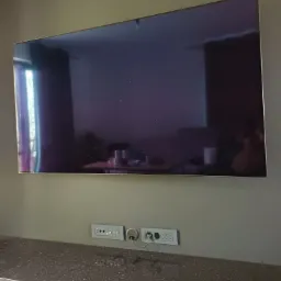 Telewizor LCD zawieszony na ścianie nad blatem z gniazdkami elektrycznymi i telekomunikacyjnymi, widoczne kable i odbicie wnętrza w ekranie.