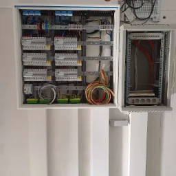 Otwarta szafa rozdzielcza z zainstalowanymi bezpiecznikami, przewodami i listwami zaciskowymi, obok szafka telekomunikacyjna z patch panelem i listwą zasilającą.