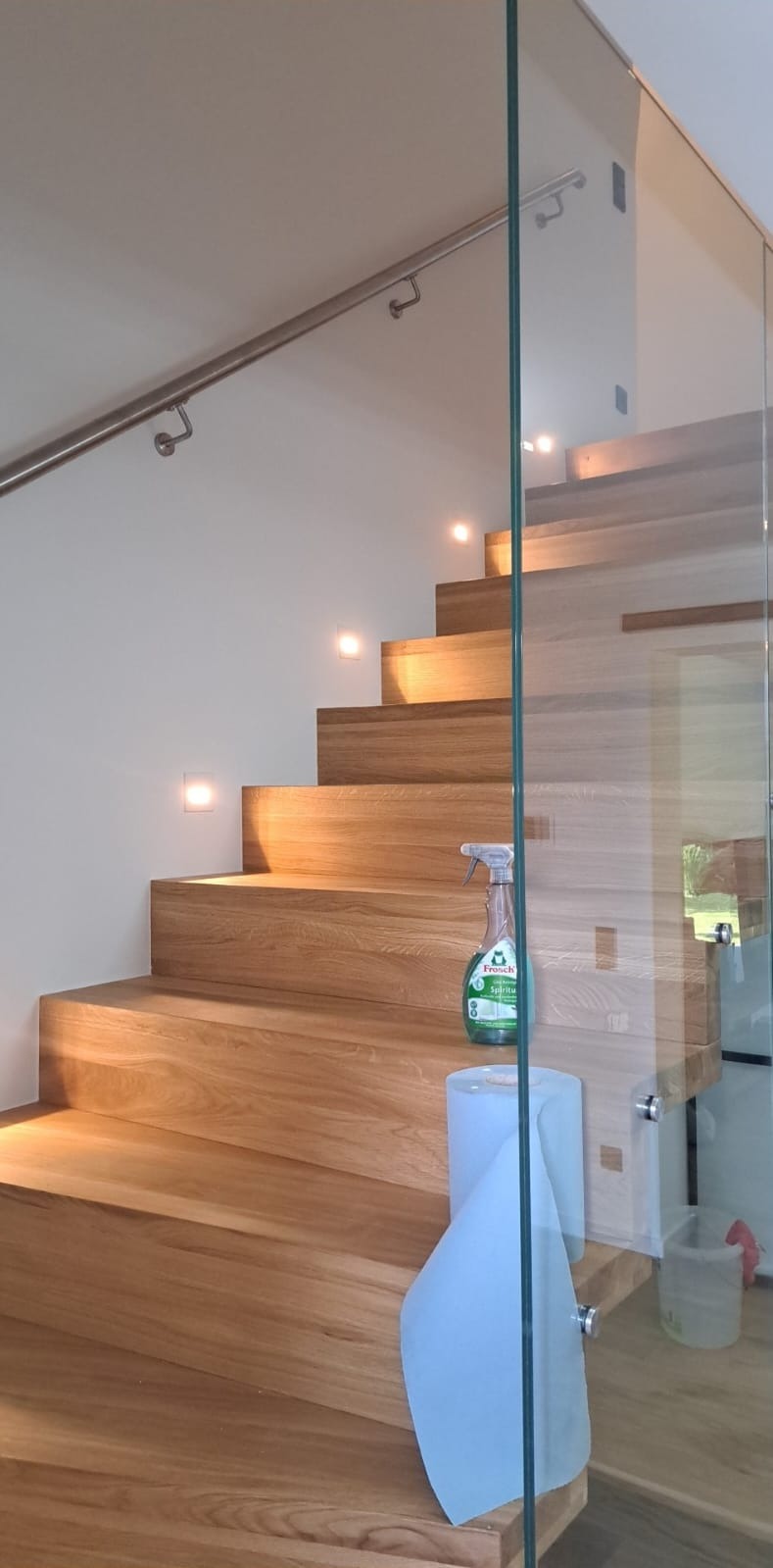 Drewniane schody z podświetleniem i szklaną balustradą w domu w Krośnie, po czyszczeniu widoczny płyn do mycia i ręcznik papierowy.
