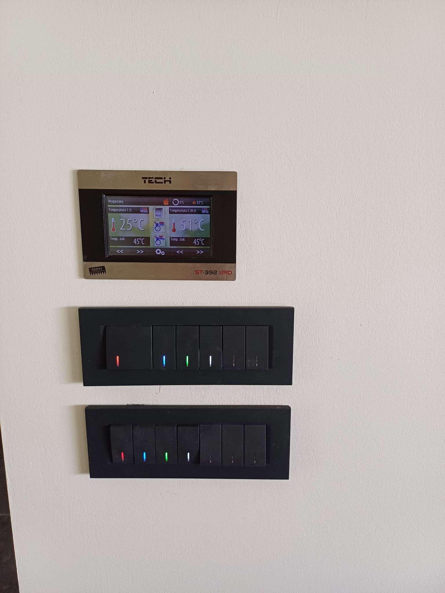 Panel sterowania ogrzewaniem TECH ST-392 zPID nad dwoma rzędami czarnych włączników światła z diodami LED na białej ścianie.