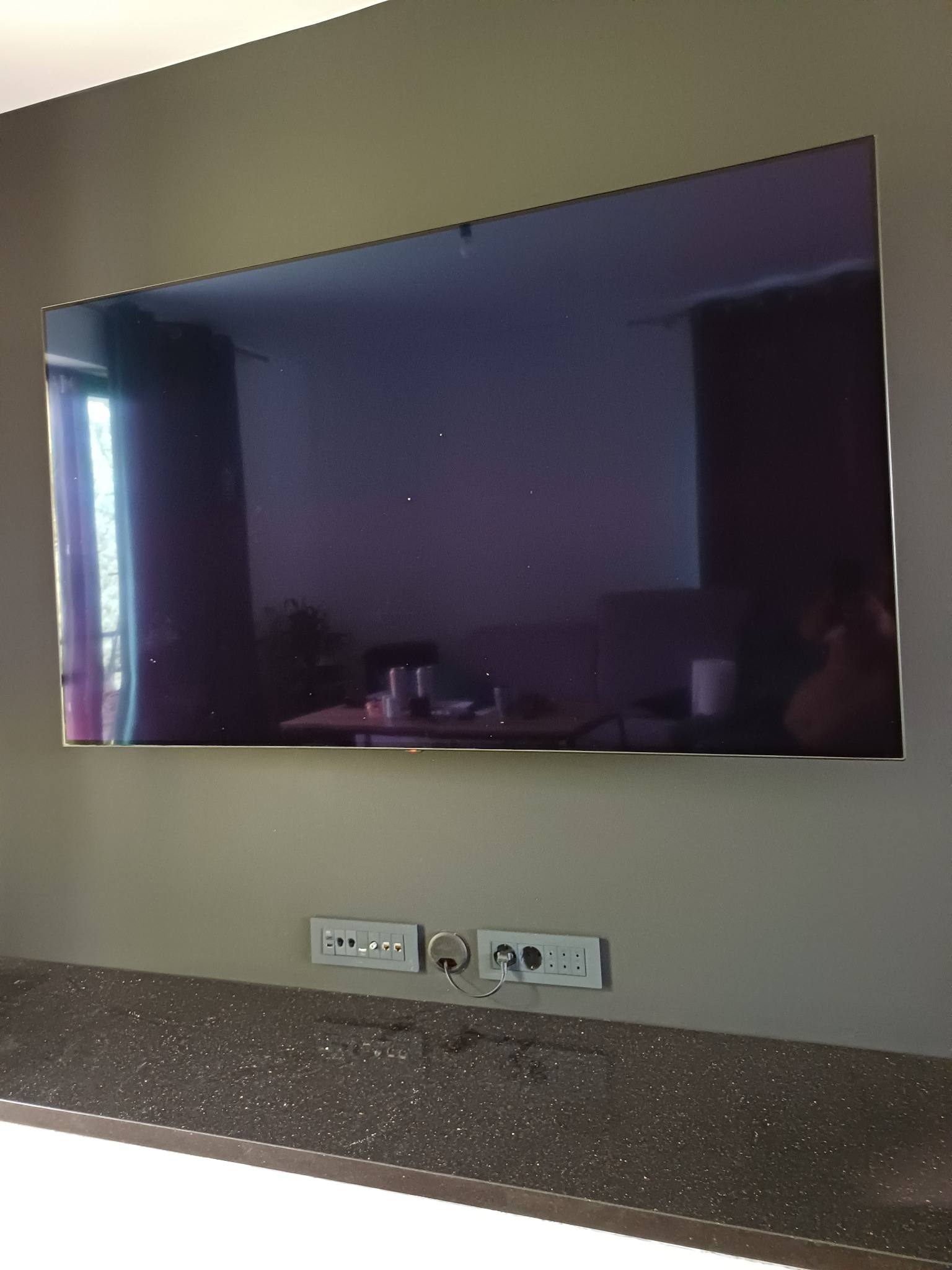 Telewizor LCD zawieszony na ścianie nad blatem z gniazdkami elektrycznymi i telekomunikacyjnymi, widoczne kable i odbicie wnętrza w ekranie.