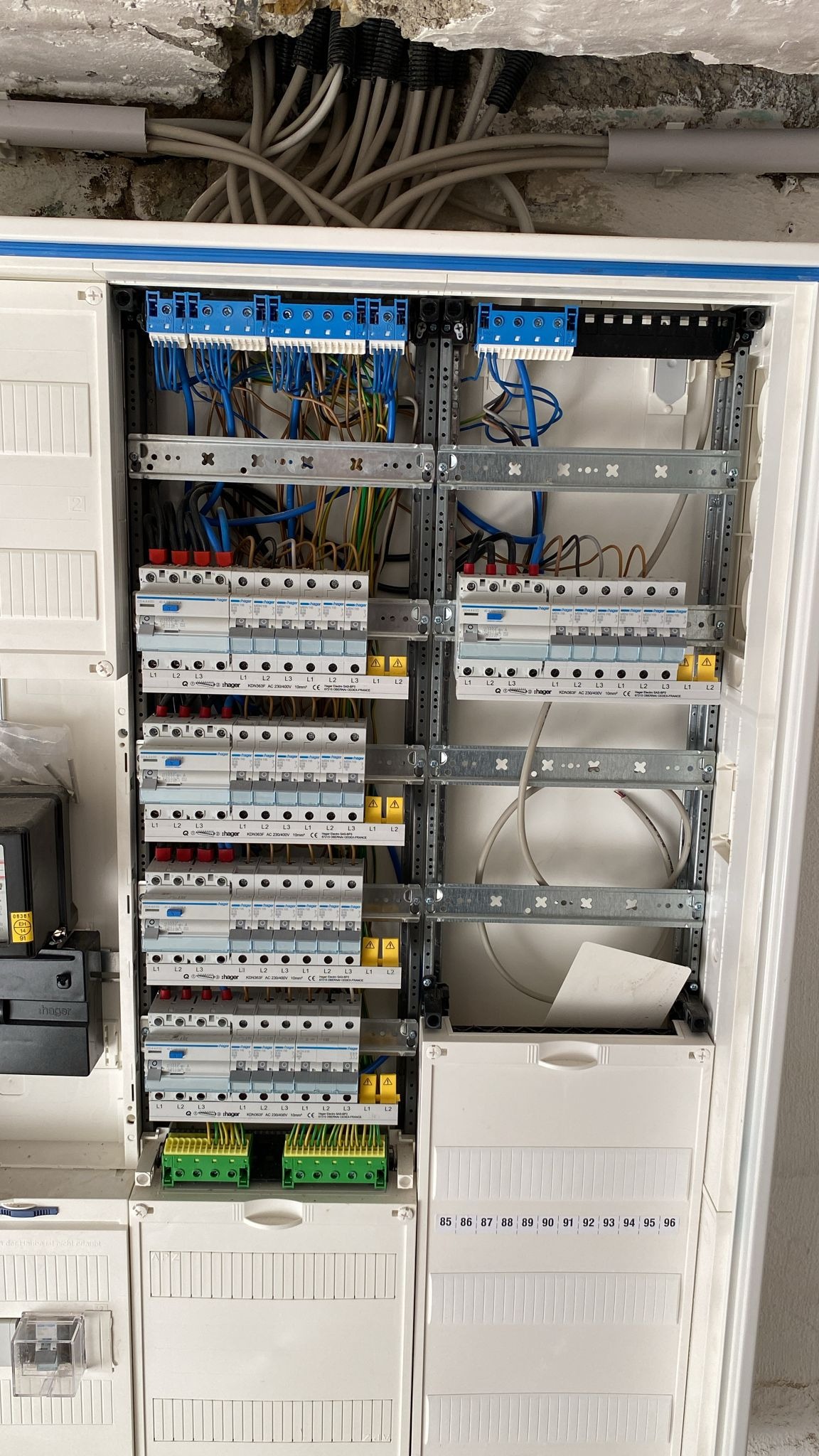 Otwarta skrzynka rozdzielcza z widocznymi zabezpieczeniami, przewodami i listwami zaciskowymi marki Hager, zamontowana na ścianie z widocznymi śladami prac remontowych.