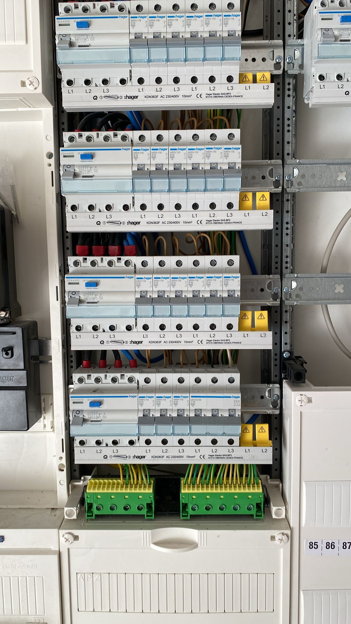 Rozdzielnia elektryczna z zamontowanymi bezpiecznikami i podłączonymi przewodami, widoczne oznaczenia L1, L2, L3 oraz żółte naklejki ostrzegawcze.