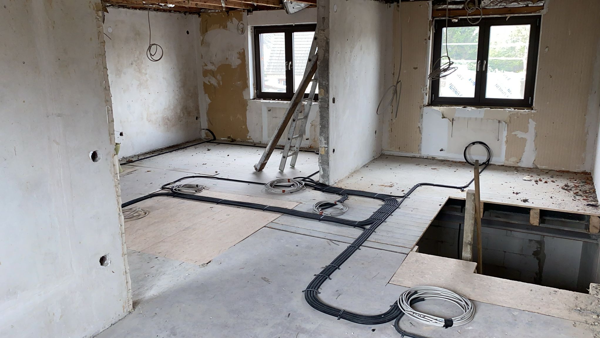 Wnętrze w trakcie remontu z widocznymi kablami elektrycznymi ułożonymi na podłodze, drabina oparta o ścianę, ślady po starych tapetach na ścianach, częściowo odsłonięta podłoga.