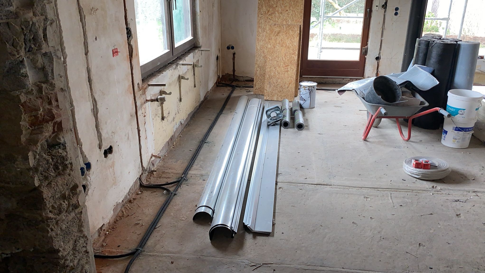 Wnętrze w trakcie remontu: widoczne bruzdy w ścianie pod instalacje elektryczne, rury, kable, taczka z gruzem i materiałami budowlanymi, okno z widocznymi mocowaniami grzejników.