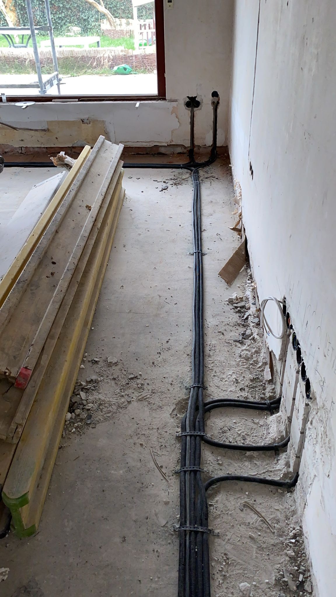 Wnętrze w trakcie remontu z widocznymi przewodami elektrycznymi w peszlach, mocowanymi do ściany, oraz przygotowanymi otworami pod gniazdka elektryczne. Obok leżą drewniane deski.