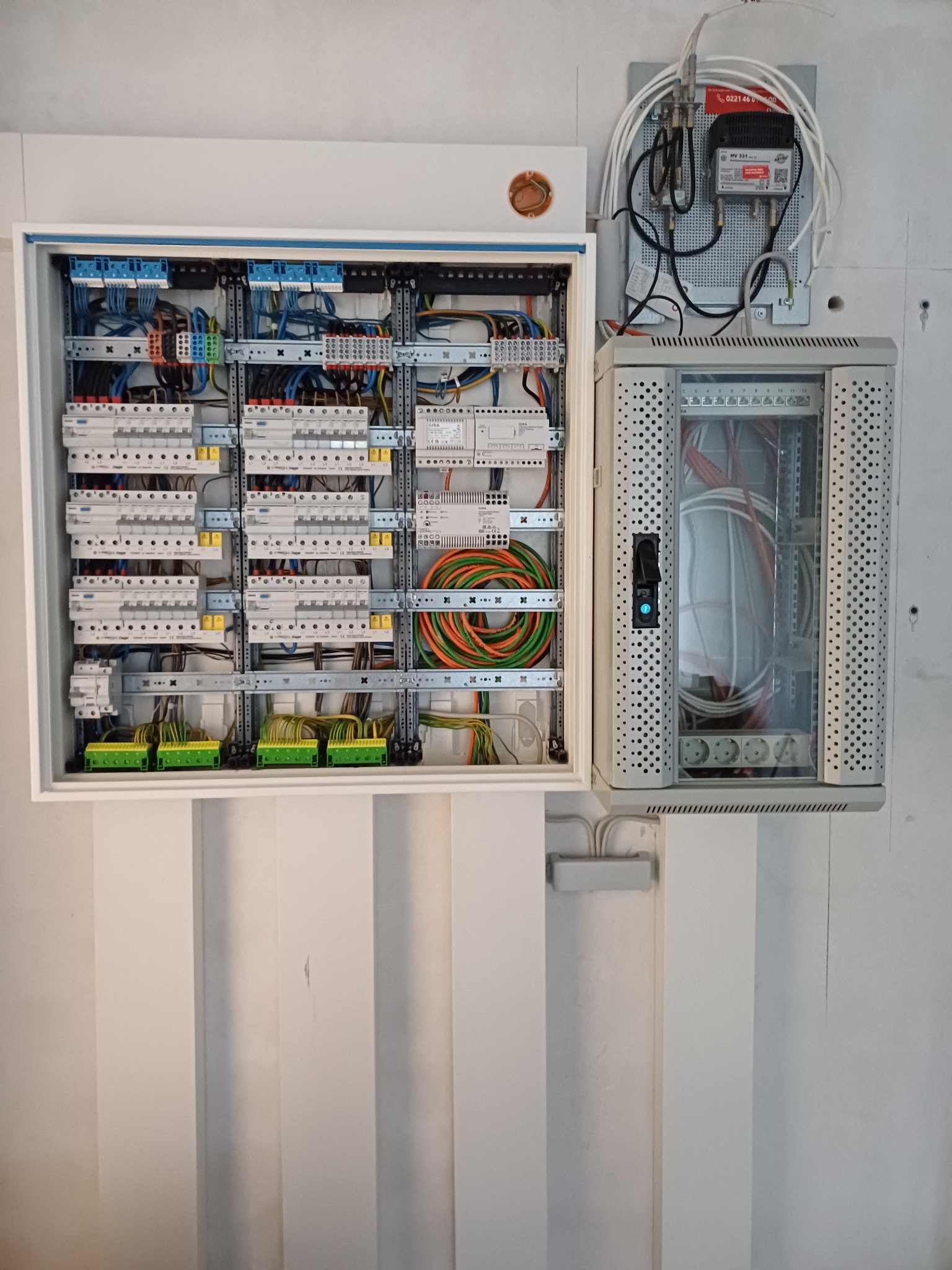 Otwarta skrzynka rozdzielcza z widocznymi bezpiecznikami, przewodami i złączkami, obok szafka telekomunikacyjna z perforowanymi ściankami i gniazdami zasilającymi, zamontowane na białej ścianie.
