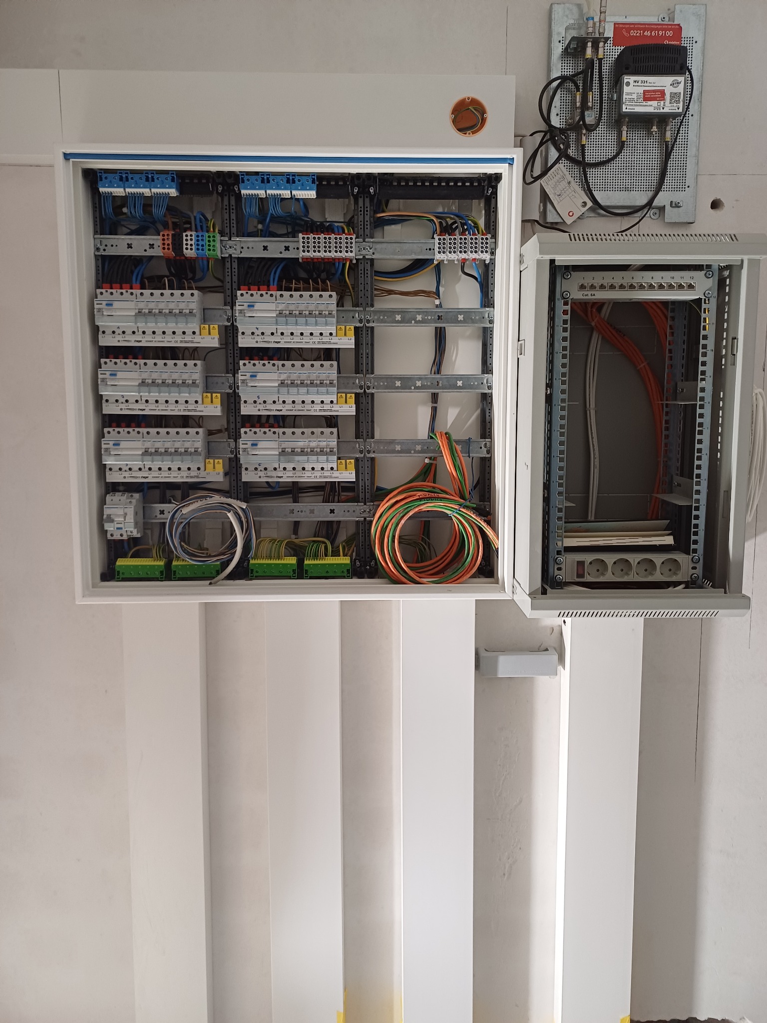 Otwarta szafa rozdzielcza z zainstalowanymi bezpiecznikami, przewodami i listwami zaciskowymi, obok szafka telekomunikacyjna z patch panelem i listwą zasilającą.