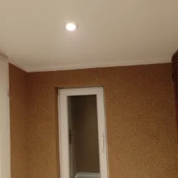 Wnętrze pokoju z oknem PCV, wykończonego brązową tapetą strukturalną, widoczna lampa sufitowa, białe listwy przypodłogowe i fragment drewnianej szafy