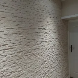 Ściana wykończona dekoracyjną okładziną imitującą kamień, z widocznym fragmentem białych drzwi z ciemną klamką i fragmentem szarej podłogi.