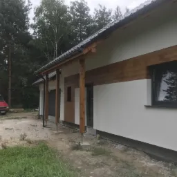 Nowoczesny dom jednorodzinny z białą elewacją i drewnianymi akcentami, widoczny czerwony samochód dostawczy na podwórzu, w tle las.