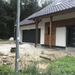 Nowo wybudowany dom jednorodzinny z elementami drewnianymi na elewacji, widoczny garaż z szarą bramą, teren wokół domu w trakcie prac wykończeniowych, na pierwszym planie słup oświetleniowy.