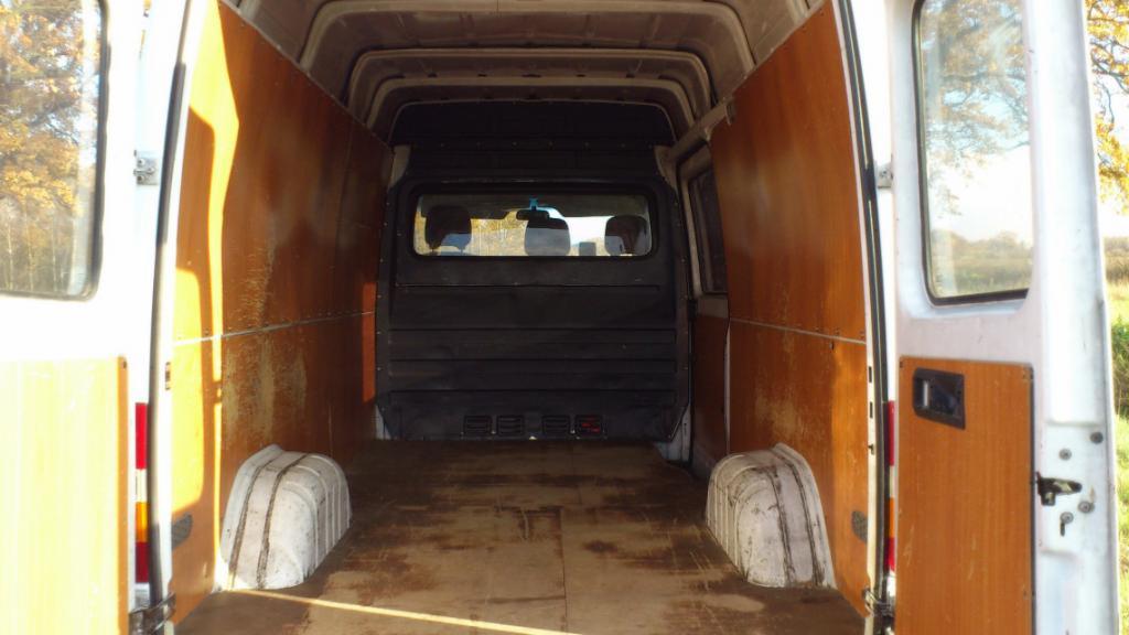 Wnętrze brudnego furgonetki z drewnianą podłogą i ścianami, widok z tyłu na otwarte drzwi i otoczenie drzew.