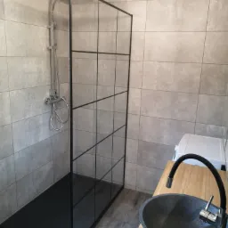 Nowoczesna łazienka z kabiną prysznicową typu walk-in z czarnymi profilami, umywalką nablatową w kolorze grafitowym i baterią z elastyczną wylewką na blacie z jasnego drewna.