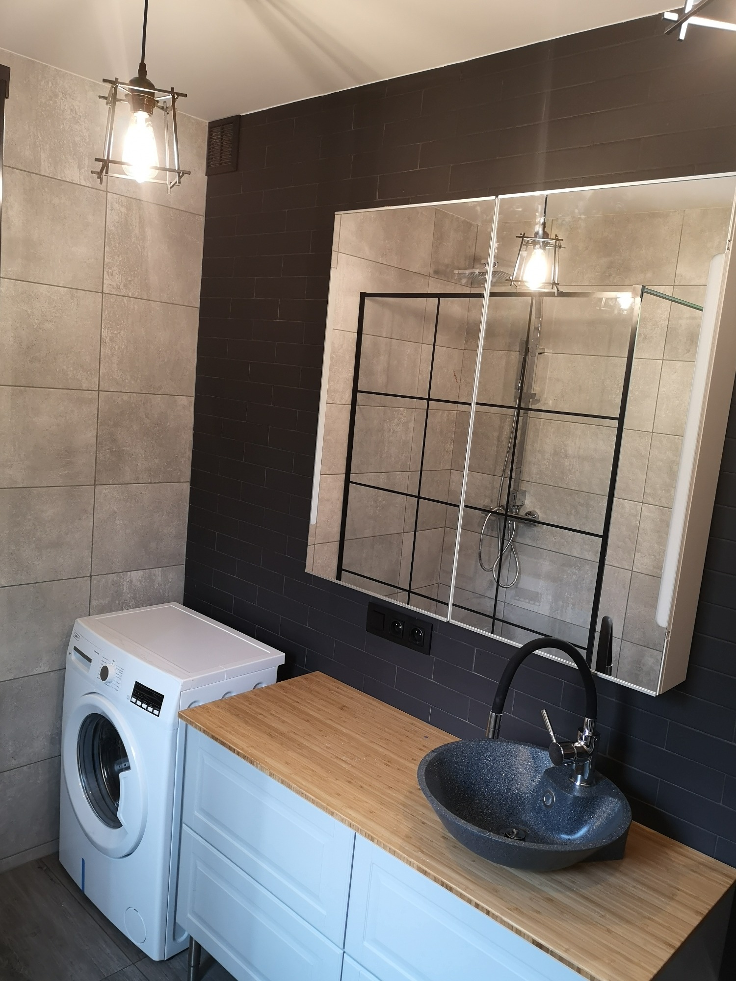 Nowoczesna łazienka z szarą armaturą, umywalką z czarnego kamienia na blacie z jasnego drewna, pralką obok szafki, czarnymi płytkami na ścianie i lustrem z czarnymi szprosami odbijającym kabinę...