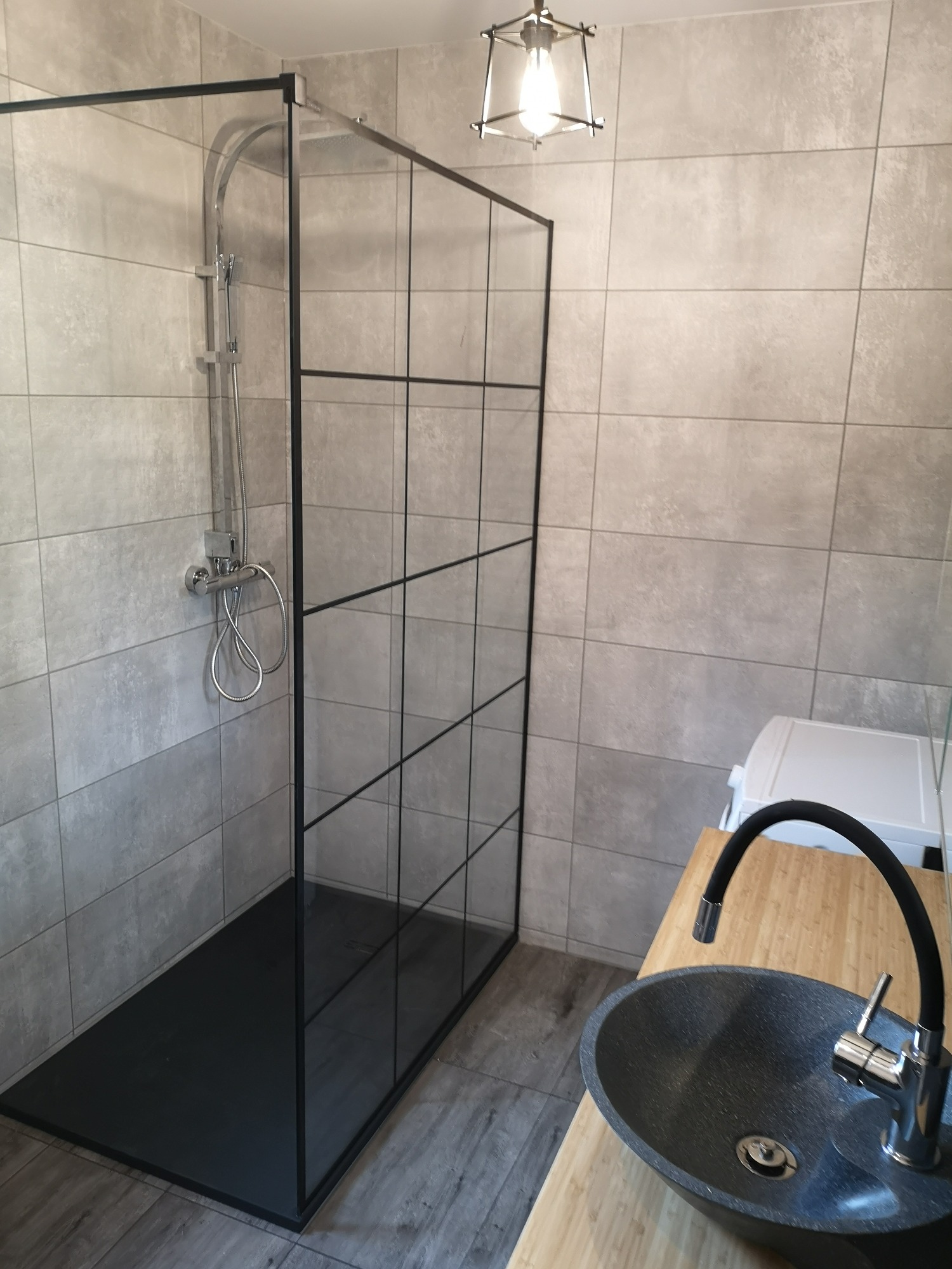 Nowoczesna łazienka z kabiną prysznicową typu walk-in z czarnymi profilami, umywalką nablatową w kolorze grafitowym i baterią z elastyczną wylewką na blacie z jasnego drewna.