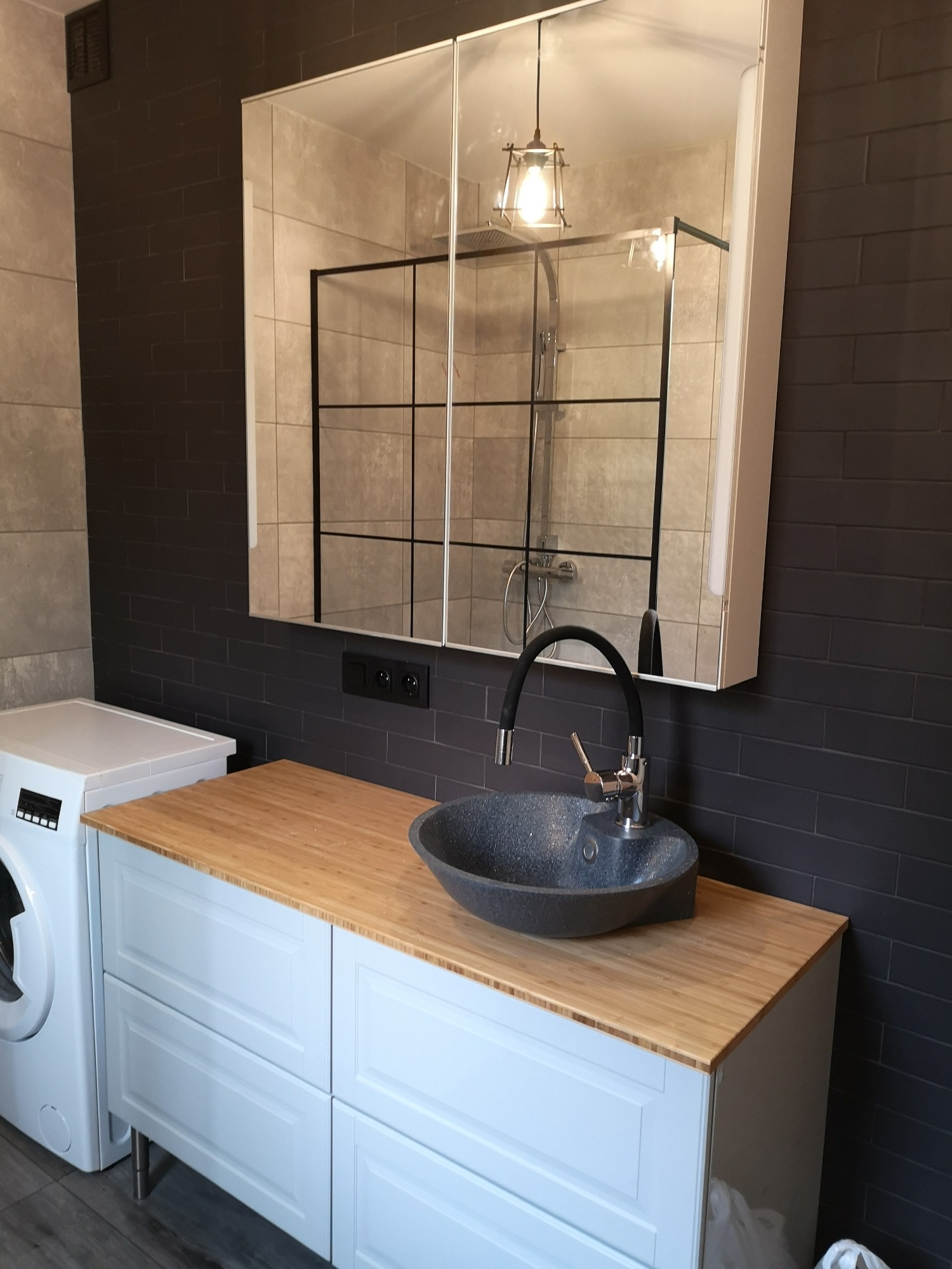 Nowoczesna łazienka z umywalką w odcieniach szarości, drewnianym blatem, białymi szafkami z czterema szufladami i pralką obok. W tle ciemnoszare płytki imitujące cegłę i lustro z odbiciem kabiny...