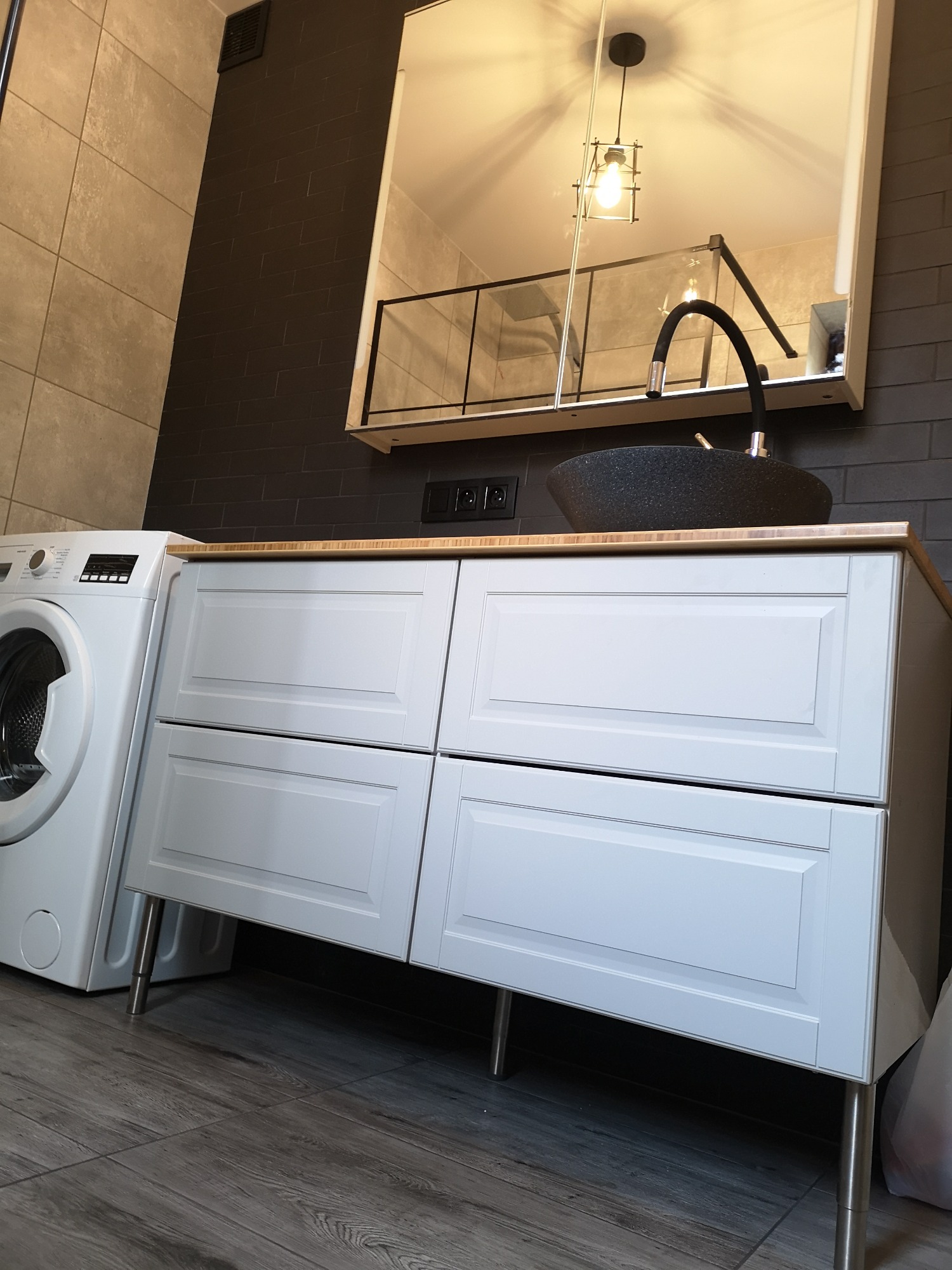 Nowoczesna łazienka z białą szafką pod umywalkę na metalowych nóżkach, czarną umywalką w kształcie misy, baterią w kształcie łuku i pralką obok, wykończona płytkami w odcieniach szarości i czerni.