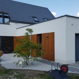 Nowoczesny dom z białymi ścianami, grafitowym dachem i drewnianymi elementami elewacji, z widocznym tarasem wyłożonym jasnymi płytami i ozdobnym drzewkiem na tle żwirowego podłoża.