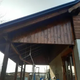 Drewniana altana z bejcowanymi deskami, widoczne podpory, zadaszenie i fragment ściany budynku z oknem przesuwnym, w tle drzewa bez liści.