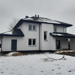 Niedawno otynkowany dom jednorodzinny z czarną dachówką i panelami słonecznymi, częściowo pokryty śniegiem, z widocznymi śladami prac ziemnych i stertą piasku przy wejściu.