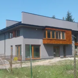 Nowoczesny dom z szarą elewacją i drewnianym wykuszem, widoczne okna z drewnianą stolarką, w tle iglaste drzewa, ogrodzenie z siatki.