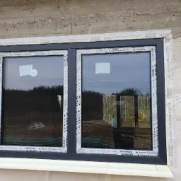 Zamontowane, dwuskrzydłowe okno z czarną ramą, zabezpieczone taśmą uszczelniającą na surowej ścianie budynku w trakcie budowy. Widoczne refleksy drzew w szybach.